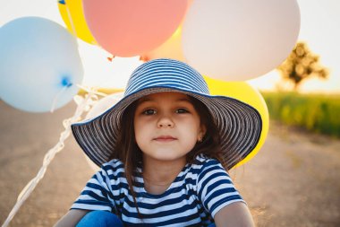 Little girl sitting on road with colorful balloons in hands at sunset.