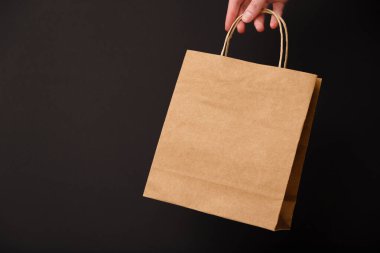 Female hand holding brown craft blank shopping bag isolated on black background. Black friday sale, discount, recycling, shopping and ecology concept