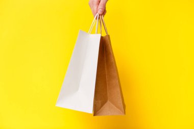 Female hand holding two shopping bags isolated on yellow background. White and brown craft blank paper bags in hand. Black friday sale, discount, shopping and ecology concept