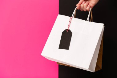 Female hand holding two shopping bags with price tag isolated on pink and black background. White and brown craft blank paper bags in hand. Black friday sale, discount, shopping and ecology concept