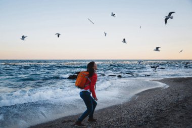 Turuncu sırt çantalı mutlu kız deniz kenarında akşam vakti martıları besliyor.