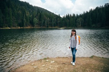 Young girl, tourist with orange backpack standing on the bank of the big mountain lake surrounded by forest. Travel destination concept.