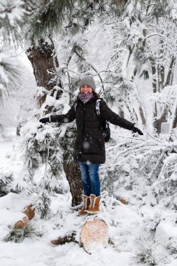 Smiling young woman standing on fallen tree after sleet load and snow in a snow-covered winter park. Girl enjoying snowy winter, frosty day. Walk in winter forest.