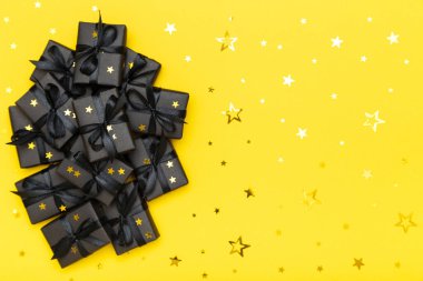 Black gift boxes with black ribbons and golden glitter stars confetti sparkles on bright rich yellow background. Black Friday sale flat lay, top view, copy space. Holiday, festive, party backdrop