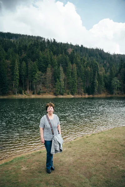 Senior beautiful woman standing on the bank of the mountain lake surrounded by forest. Travel concept.