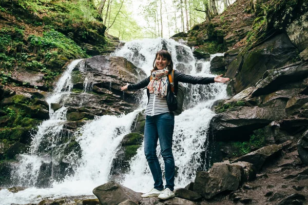 Happy smiling tourist girl with backpack at cold fresh waterfall. Waterfall Shypit, cascade in Pylypets in forest. Carpathian Mountains, Ukraine