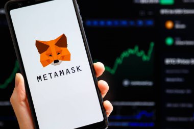 Ukraine, Odessa - October, 9 2021: Hand holding mobile with MetaMask app running at smartphone screen with trading candlestick chart at background. MetaMask is software crypto wallet