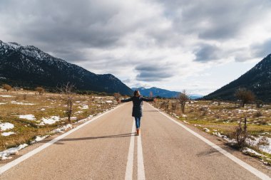 Back view of young woman walking along empty asphalt road in the middle of mountainous area with raised hands. Highlands of Greece, winter time. Freedom, travel concept.