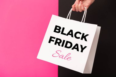 Female hand holding white shopping paper bag with Black Friday Sale text on pink and black background. Sale, discount, shopping concept