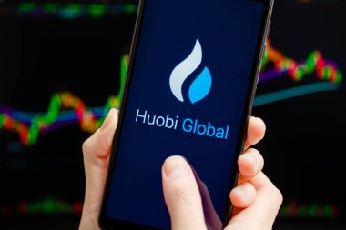 Ukraine, Odessa - October, 9 2021: Hands holding mobile with Huobi Global app running at smartphone screen, trading candlestick chart at background. Huobi - cryptocurrency exchange, trading platform