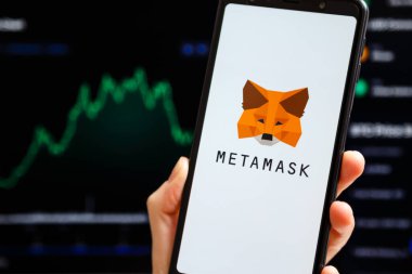 Ukraine, Odessa - October, 9 2021: Hand holding mobile with MetaMask app running at smartphone screen with trading candlestick chart at background. MetaMask is software crypto wallet
