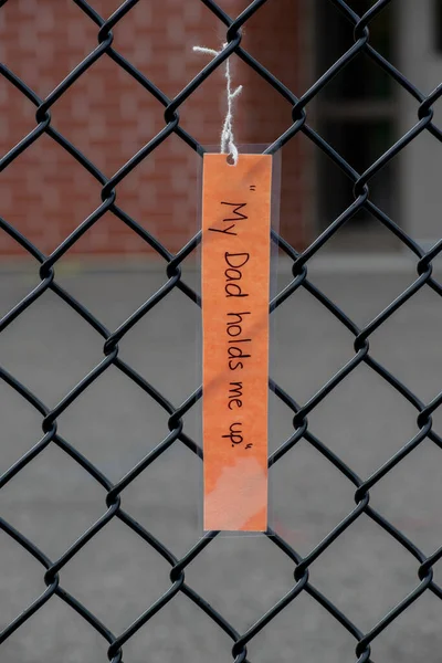 A laminated note card (my dad holds me up)is hanging from a chain link fence, Ontario, Canada - stock photography