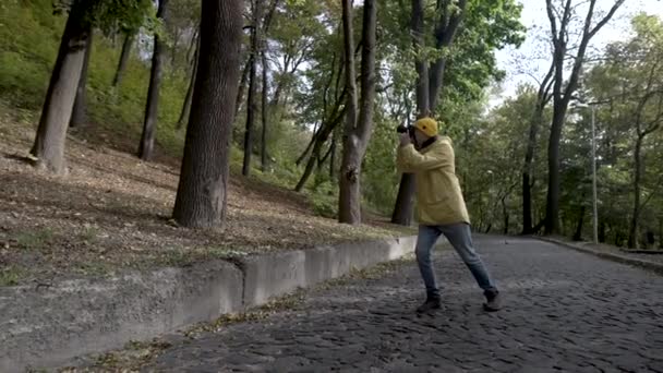 Utazási fotós csinál egy képet a parkban fák. Egy sárga köpenyes fiatalember sétál végig az úton. Turista digitális fényképezőgép. A siker öröme — Stock videók