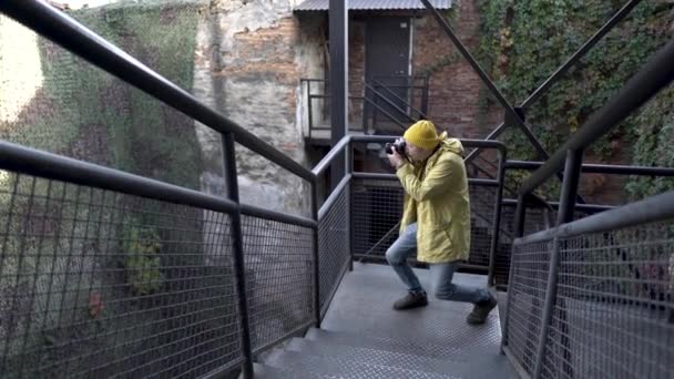 Cestovní fotograf fotí, jak stojí na kovové konstrukci. Mladý muž ve žlutém plášti. Turistické a digitální fotoaparáty. — Stock video