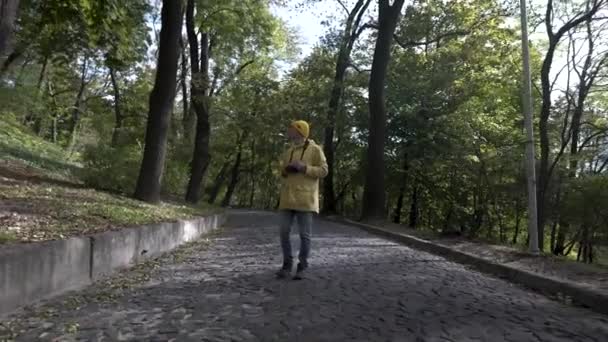 Fotograf podróżny robi zdjęcie w parku z drzewami. Młody mężczyzna w żółtym płaszczu idzie ulicą. Cyfrowa kamera turystyczna. Radość sukcesu — Wideo stockowe