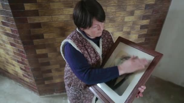 Elderly woman becoming sad while holding and looking at old photo portrait of ancestor. Old lady looking upset and having nostalgia while standing at home. Concept of feelings, emotions and memories — Vídeo de stock