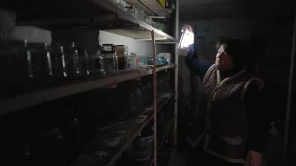 An elderly woman looking for canning on the shelves in a dark old cellar using a lantern. Rural life. Survival of poor people in difficult times. A peasant woman in an old clothes. — Vídeo de stock