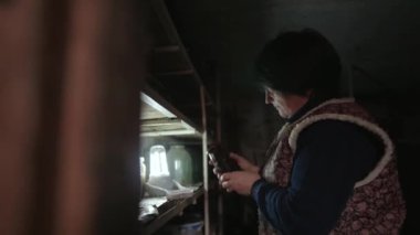 Typical rural life. An elderly woman is sorting through jars of homemade pickled vegetables on the shelves in a dark old cellar. Peasant woman in old clothes. Preparing for winter