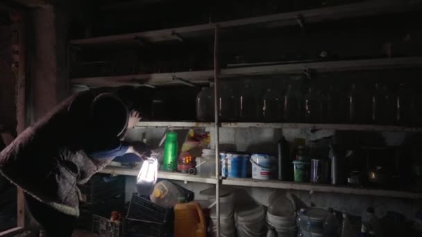 Пожилая женщина ищет консервы на полках в темном старом подвале, используя фонарь. Сельская жизнь. Выживание бедных людей в трудные времена. Крестьянка в старой одежде. — стоковое видео