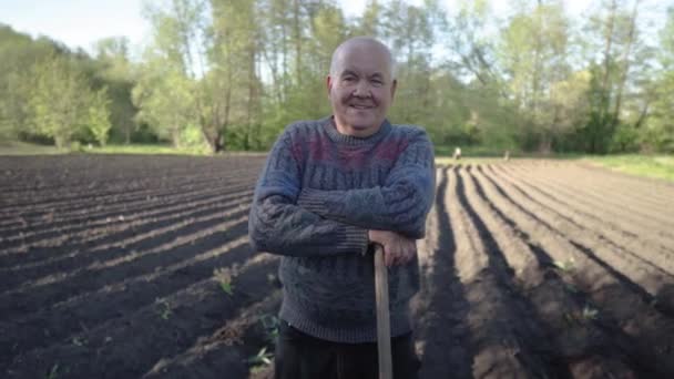 Portrait of a happy elderly farmer after working in the field. The peasant poses in front of the camera. The villager standing on the field with hoe. High-quality cultivated land in the background — стоковое видео