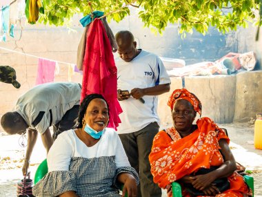 Bour, Senegal - Aralık 2021 civarı. Dışarıdaki plastik sandalyede oturan renkli geleneksel elbiseli kimliği belirsiz kadınlar. Köyde aile hayatı. Afrika 'da yoksulluk