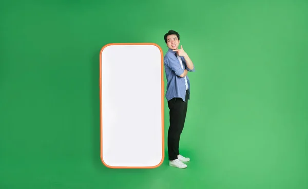 青の背景に隔離された大きなスマートフォンの隣に立つアジア系男性の全身像 — ストック写真