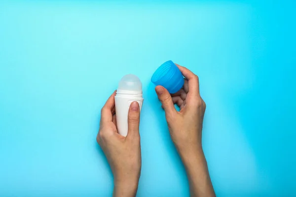woman hand holding and showing white bottle of armpit deodorant on blue background. Female daily beauty product. Closeup. Empty place for text or logo.