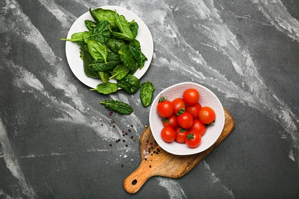 spinach leaves in a plate with tomatoes in a bowl on a wooden cutting board with salt and pepper. Black background. top view