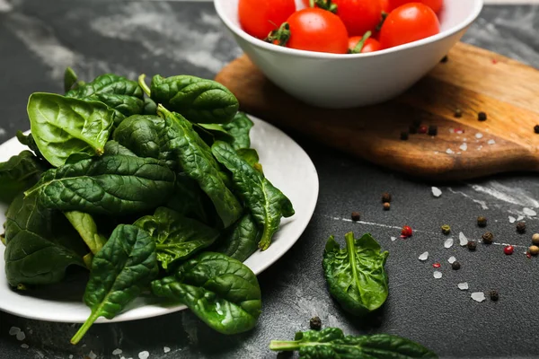 spinach leaves in a plate with tomatoes in a bowl on a wooden cutting board with salt and pepper. Black background