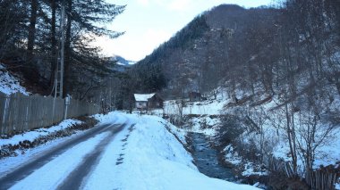 Snowy rural landscape in Ciungetu, Romania. A half frozen river flows by the road and along the spruce forest, to complete this winter countryside landscape.