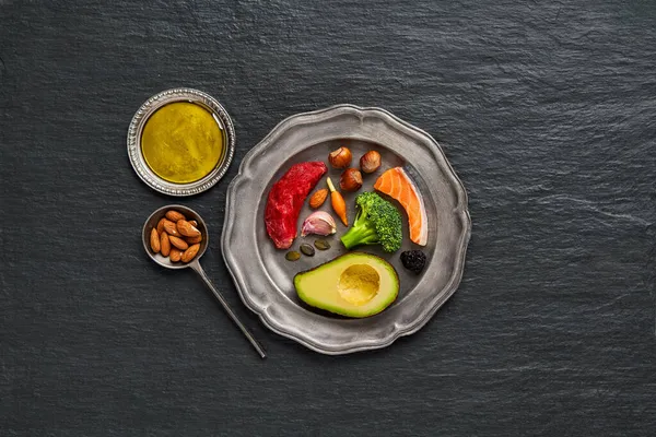  Ketojenik diyet yiyeceğiyle kaplı bir tabak. Sığır eti, somon, yumurta, brokoli, domates, fındık, havuç, mantar, salatalık, hurma. Fotokopi alanı olan karanlık bir arka planda diyet yemekleri. Üst Manzara. Düz yatıyordu