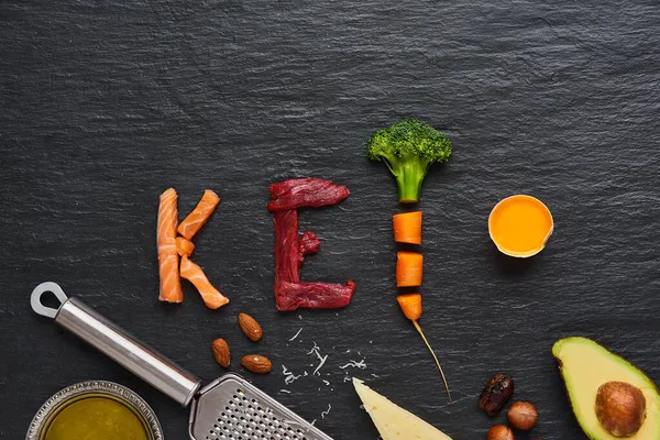 Ketojenik gıdalardan yapılmış keto kelimesi, yeni yıl sağlık diyeti çözeltisi sığır eti, somon, yumurta, brokoli, domates, fındık, havuç, mantar, salatalık, hurma. Fotokopi alanı olan koyu arkaplanda diyet yemekleri.            