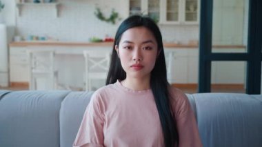 Evdeki kanepede oturan kendine güvenen genç Asyalı bir kadının portresi. Çekici genç Asyalı etnik kadın kameraya özgüvenle bakıyor, evdeki kanepede oturuyor.