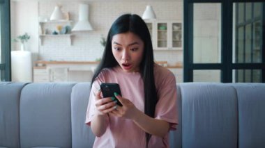 Genç ve çekici Asyalı etnik kadın oturma odasında kanepede oturuyor ve akıllı telefona yazıyor, seviniyor ve yumruğunu sıkıyor, aniden cep telefonu ekranından harika haberler okuyor.