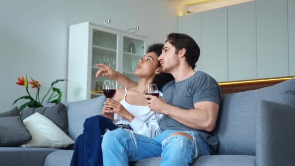 Beautiful couple sitting with glass of wine, embracing on couch, discussing, gesturing with hands, looking to the side. Happy married couple of African woman and European guy chatting together at home — Stock Video