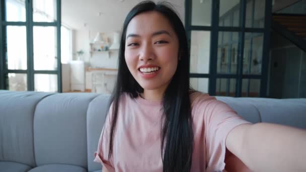 Sorrindo jovem asiático mulher fazendo selfie ou vídeo chamada enquanto sentado em casa no sofá. Retrato de blogueira fazendo um bate-papo em vídeo de seu apartamento. Ponto de vista. — Vídeo de Stock