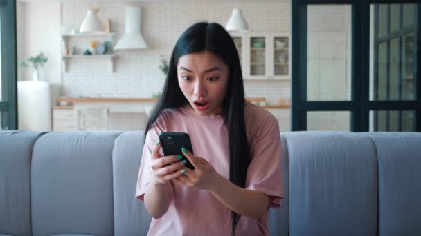 Młoda atrakcyjna azjatycka etniczka siedząca na kanapie w salonie i wpisująca się w smartfona, radująca się i zaciskająca pięść, nagle czytająca świetne wiadomości na ekranie telefonu komórkowego — Wideo stockowe