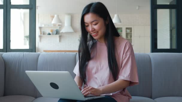 Close-up of pretty young woman of Asian ethnicity, freelancer, student focused on typing text on laptop, working remotely while sitting on sofa in living room. Distant work and e-learning concept — Stock Video