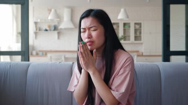 Молода жінка азіатського походження плаче, сидячи на дивані в студії, закриває обличчя долонями, нервово і тривожно озирається. Емоції засмученої жінки. — стокове відео
