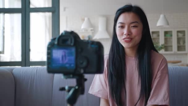 Крупный план молодого блогера, записывающего видео на камеру для подписчиков. Дружелюбная милая азиатка записывает видео с камерой, установленной на штатив, приветствие, машет рукой, представляясь. — стоковое видео