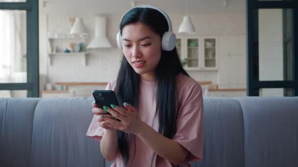 Mulher asiática bonita colocando fones de ouvido e levando seu smartphone para navegar em um aplicativo móvel e ouvir a música. Divertir-se e descansar em casa em seu dia de folga. Atividades de lazer, fim de semana — Vídeo de Stock