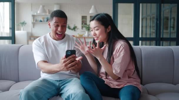 Bărbat afro-american și femeie asiatică odihnindu-se acasă, stând pe canapea, îmbrățișându-se unul pe celălalt și bucurându-se după ce au privit noi la telefon. Tineri interrasiale cuplu sentimentul de fericire împreună — Videoclip de stoc
