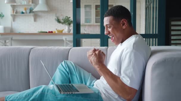 Bonito ganhador afro-americano descansando no sofá regozijando-se olhando para monitor de laptop, dançando, apertando punhos depois de receber boas notícias. Masculino celebrando sucesso aposta vitória em casa no sofá — Vídeo de Stock