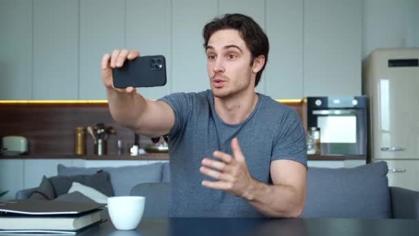 Pohledný mladý muž, který drží svůj chytrý telefon v natažené paži, dívá se na webovou kameru, zdraví svého partnera, svolává poradu přes videohovor. Online komunikační koncepce — Stock video