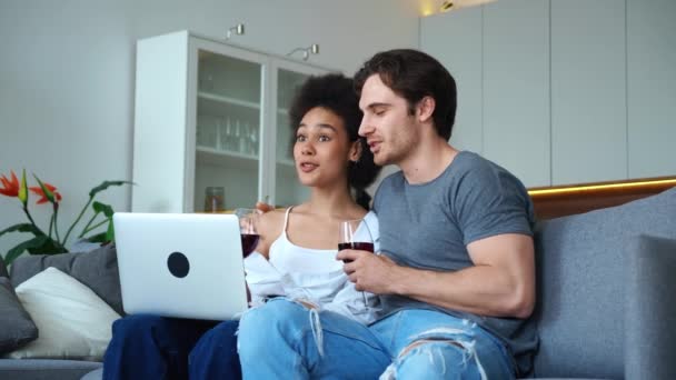 Mladý zamilovaný pár, sedící objímající se na gauči s laptopem, smějící se, radující se, flirtující se sklenicí vína v rukou. Šťastný manželský vztah, láska, náklonnost, něha — Stock video