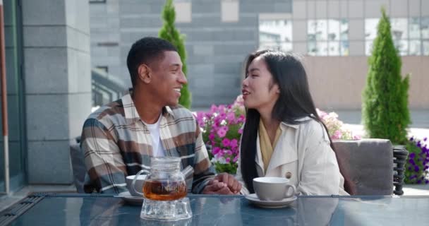 Vonzó fiatal pár ázsiai gyönyörű nő és jóképű afro-amerikai srác pihentető ül az asztalnál forró ital a nyári kávézóban, beszél, élvezi pillanatok együtt romantikus randin — Stock videók