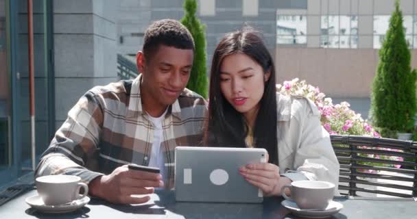 Junge multi-ethnische Paar afrikanischer Mann mit Kreditkarte und fröhliche asiatische Frau, sitzt am Tisch einer Sommerterrasse des gemütlichen Cafés, Blick auf das digitale Tablet, Online-Einkäufe tätigen — Stockvideo