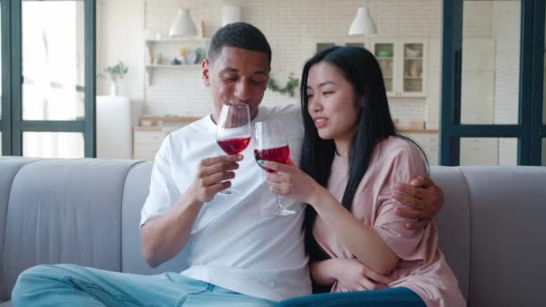 Счастливая молодая гетеросексуальная любящая пара смешанных расовых мужчин и азиатских женщин, обнимающих друг друга, сидящих на диване с бокалами красного вина и наслаждающихся счастливым временем вместе, обсуждающих, флиртующих — стоковое видео