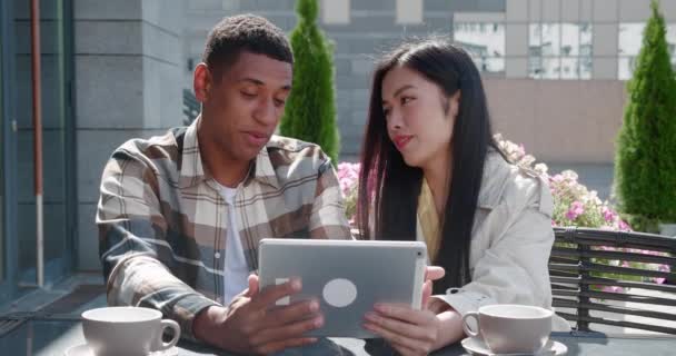 Young loving couple, two close friends sitting in the outdoor cafe on warm sunny day at romantic date, talking, looking at digital tablet in the hands of handsome guy, swiping, surfing on web pages — Stock Video