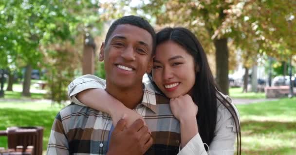 Aranyos, többnemzetiségű, szeretetteljes heteroszexuális pár, gyönyörű, boldog ázsiai nő, mosolygós, fogas mosollyal ölelgeti afro-amerikai barátját, aki gyengéden simogatja a kezét, és együtt néz a kamerába.. — Stock videók